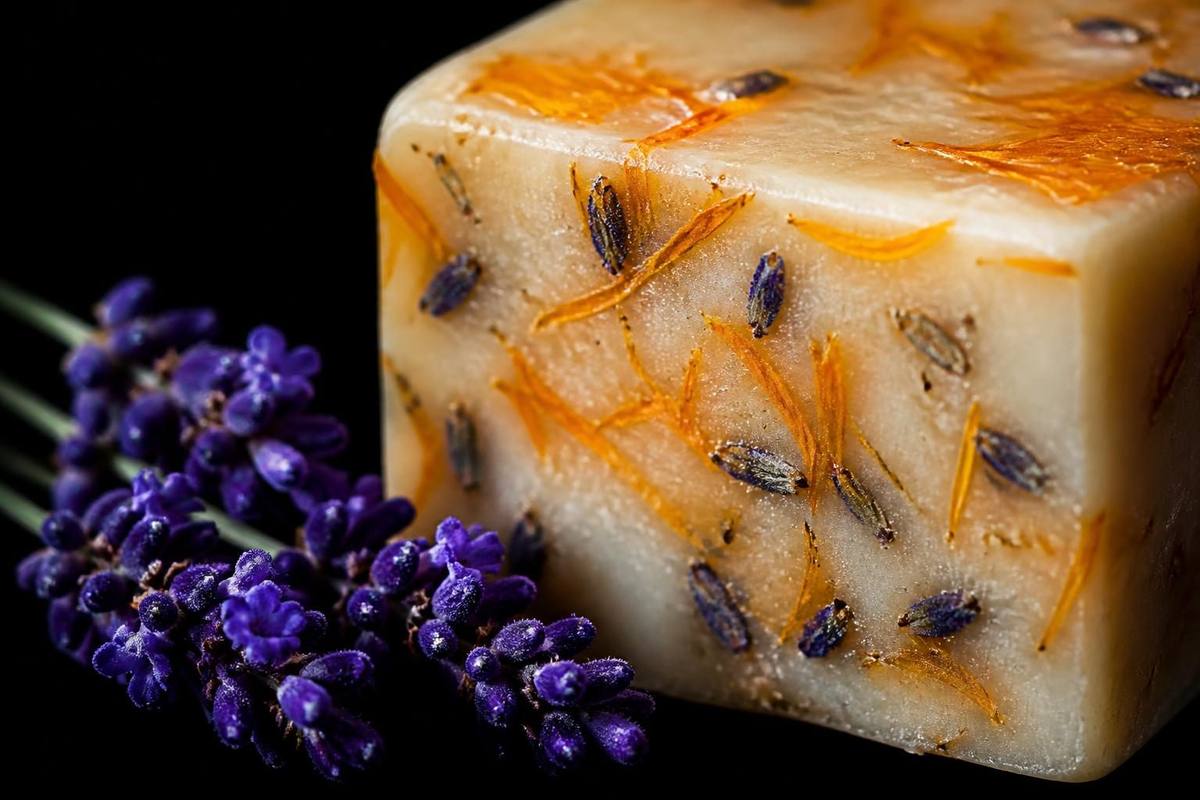Macro studio sur fond noir absolu — barre de savon à froid ivoire incrustée de pétales de calendula orangés and boutons de lavande violets, brin de lavande saturé en avant-plan.