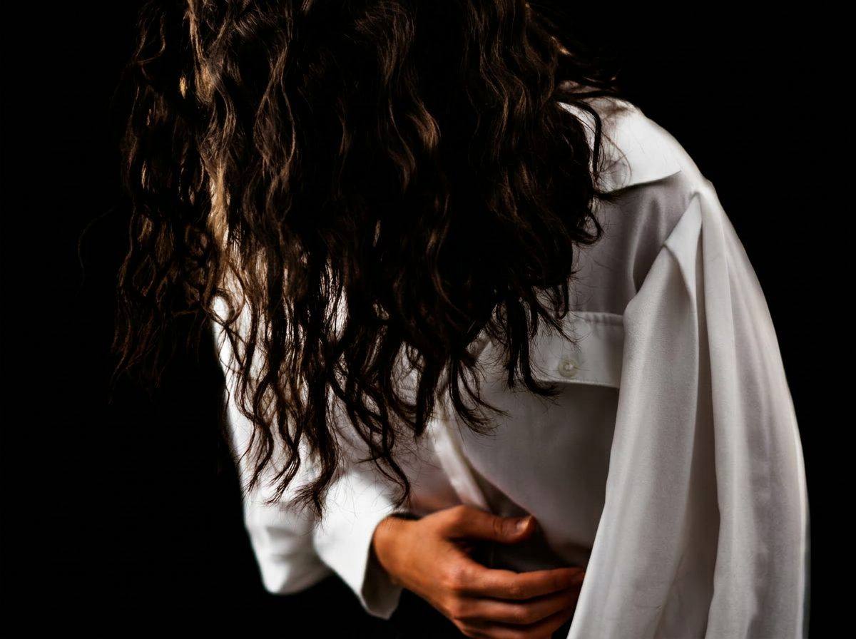Portrait noir et blanc chiaroscuro — femme aux cheveux bouclés, chemise blanche, main posée sur le ventre.